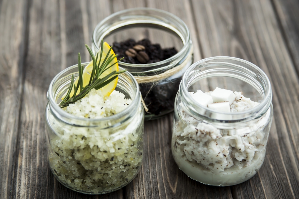 3 borcane de scrub, 2 albe , 1 inchis la culoare cu diferite plante si ingredinete, pe o masa gri