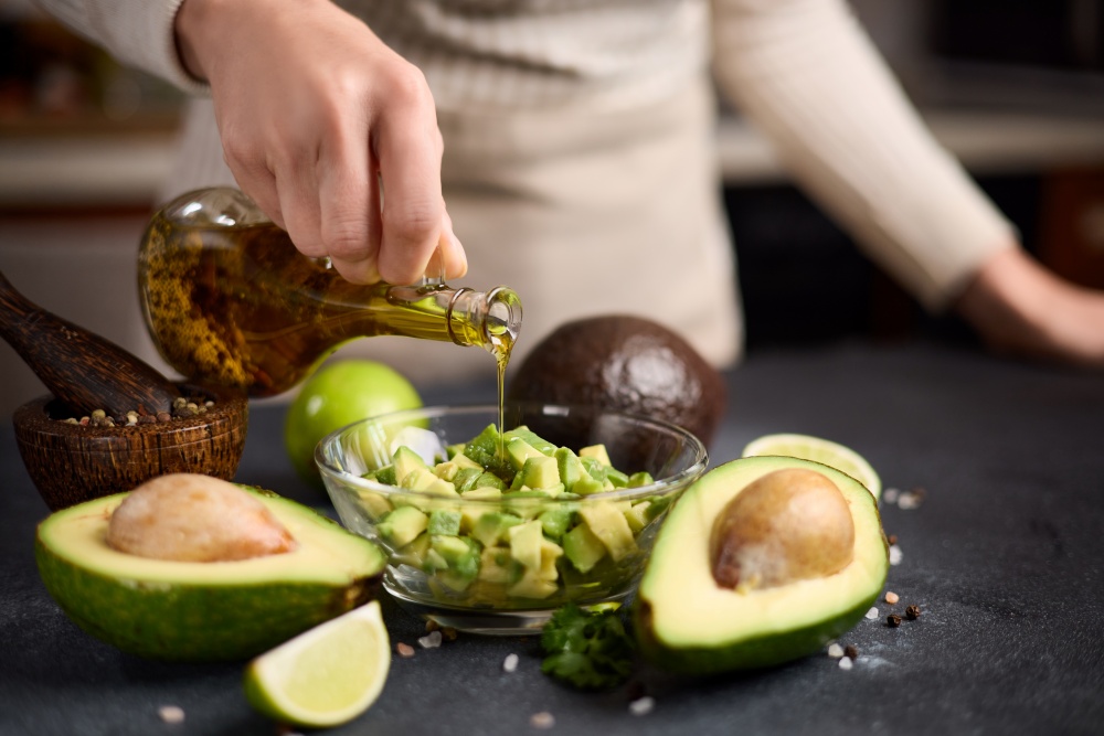 O femeie toarna ulei peste o salata de avocado. Pe langa bolul in care se afla salata se afla cateva bucati de avocado.
