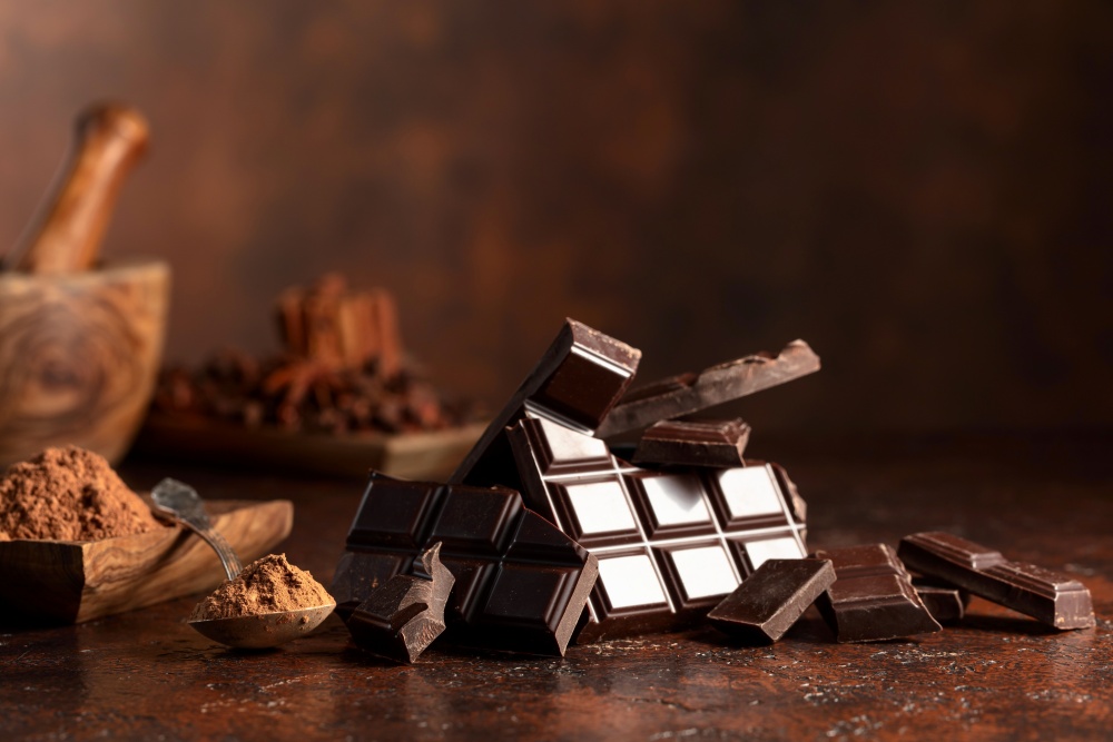 Bucati rupte de ciocolata neagra si pudra de cacao pe o masa de culoare maro.