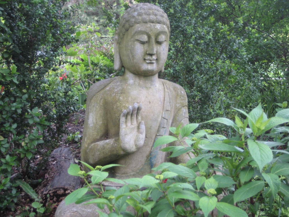 O statuie a lui Buddha intr-o gradina, tinand mana in pozitia Abhaya mudra/ mudra neinfricarii