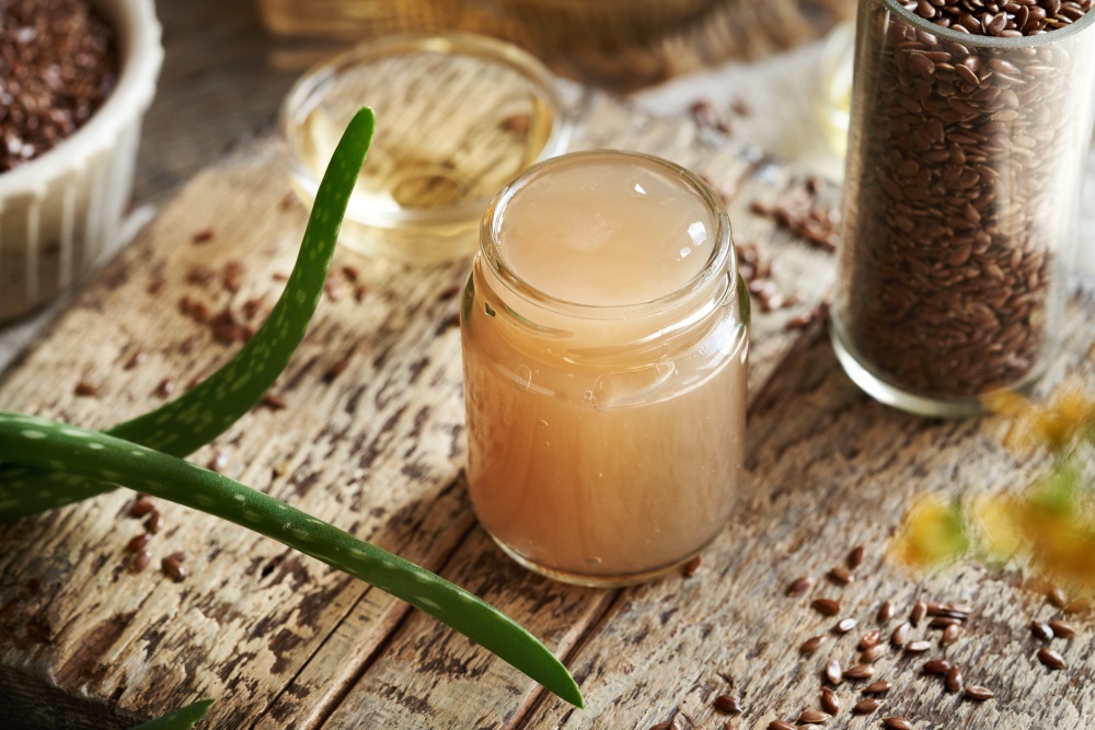O masa pe care se afla un borcansa cu gel natural de in pentru par, seminte de in intr-un recipient si o planta de aloe vera.