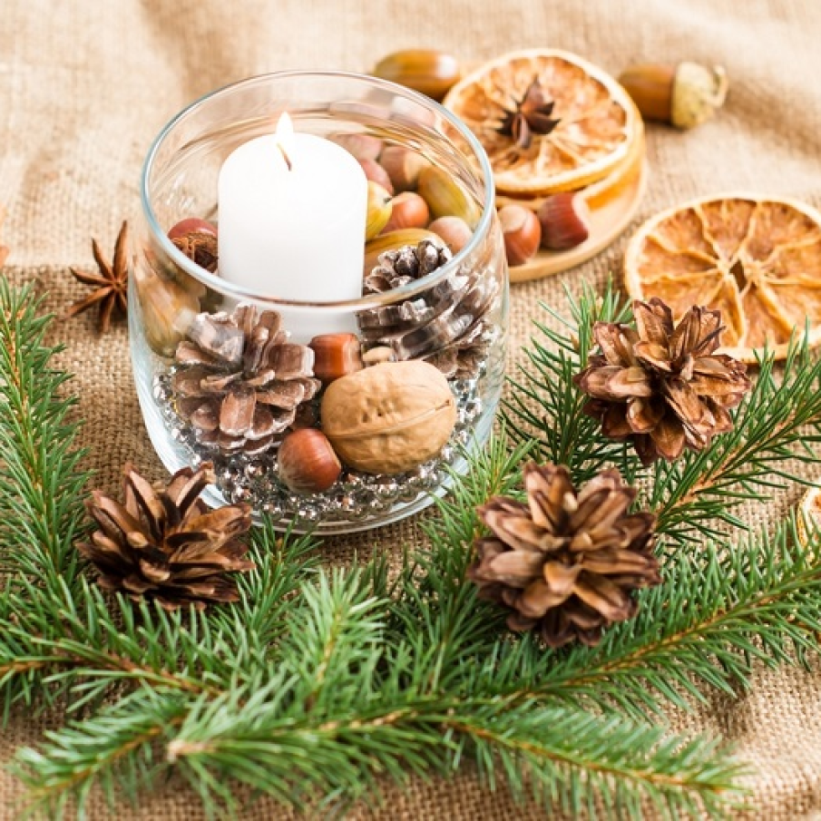 Borcan decorativ de Sărbători umplut cu fructe specifice iernii și ingrediente naturale precum nuci, ghinde, conuri și o lumânare albă 