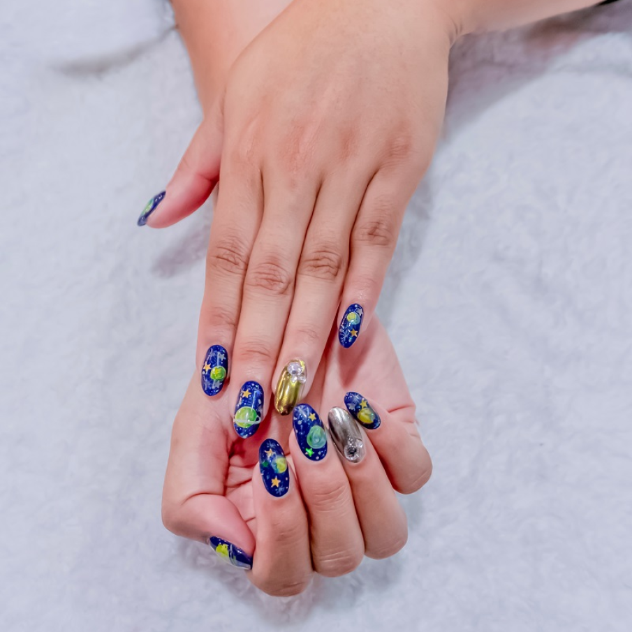 Manichiură statement cu tematică spațială. Unghiile inelarului sunt în nuanțe metalizate de auriu și argintiu, iar pe restul unghiilor regăsim motive specifice cosmosului