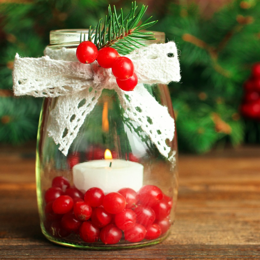 Decorațiune handmade de Crăciun: lumânare plasată într-un borcan pe fundul căruia se află fructe roșii de călin, iar borcanul este decorat cu o fundă albă din material tricotat și o crenguța de molid 
